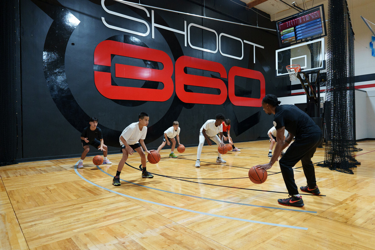 Youth basketball players practice dribbling with a coach on a Shoot 360 training court beneath a large Shoot 360 wall logo.