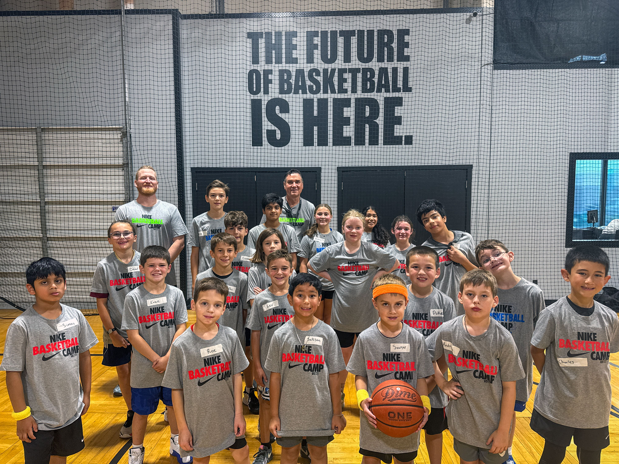A group of kids and coaches in gray Nike Basketball Camp shirts pose in a gym. A sign behind them reads THE FUTURE OF BASKETBALL IS HERE. Some kids hold basketballs and smile at the camera.